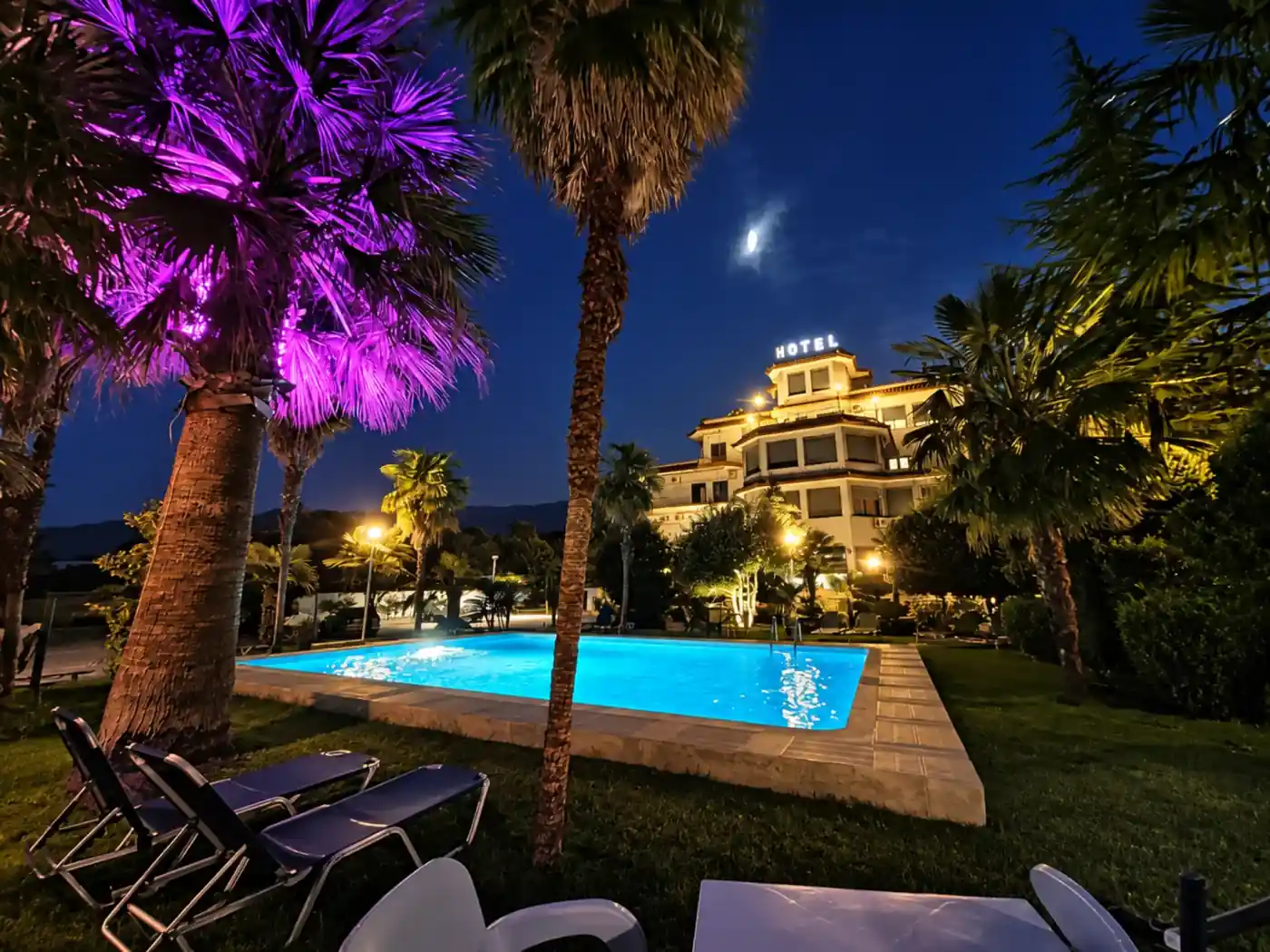 Pishina e Hotel Palma natën, e ndriçuar në ngjyrë bruzë mes palmave, me hotelin në sfond